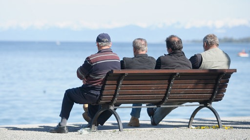 Vier Herren fortgeschrittenen Alters sitzen in Konstanz auf einer Bank am Ufer und blicken auf den Bodensee hinaus