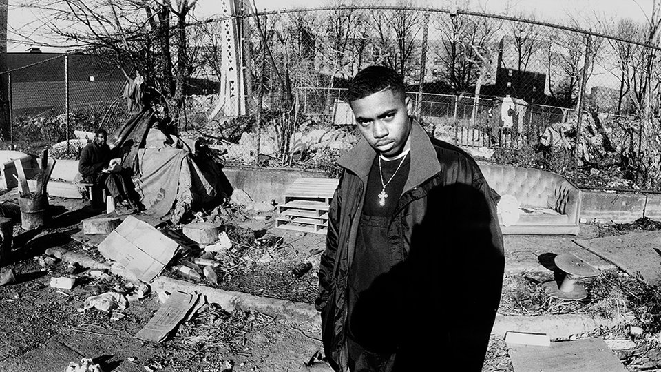 Szene aus "Nas - Time Is Illmatic": Nasir Jones vor einem zugemüllten Bürgersteig in Queensbridge; im Hintergrund ein Obdachloser auf einem Sofa